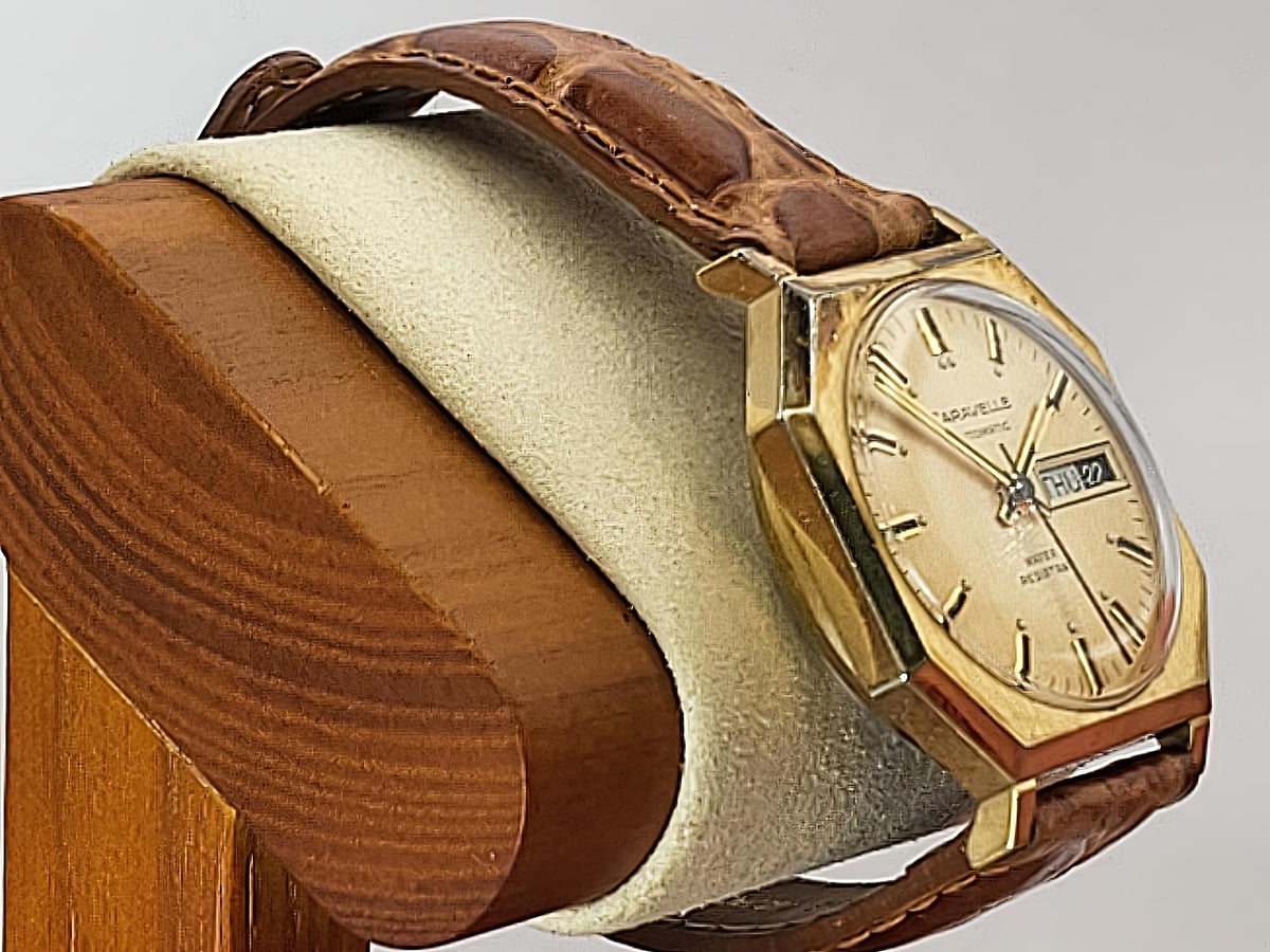A vintage Caravelle Day-Date watch with a gold-toned case, cream dial, and brown leather strap, displayed on a wooden stand with a cream velvet cushion.
