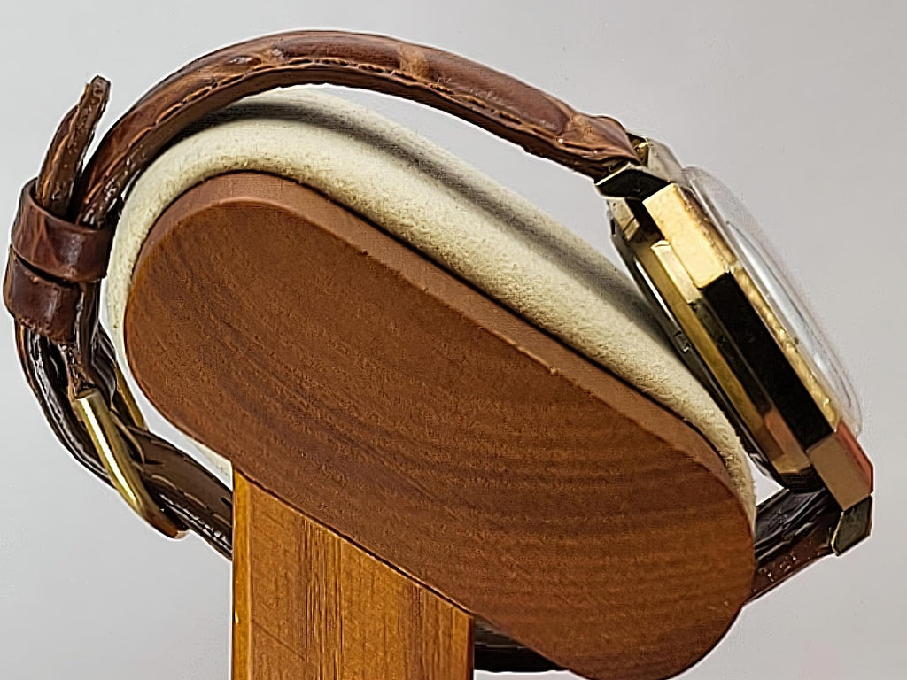 Close-up of a Caravelle Day-Date wristwatch with a brown leather strap and gold-tone case, displayed on a wooden stand against a light gray background.