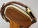 Close-up of a Caravelle Day-Date wristwatch with a brown leather strap and gold-tone case, displayed on a wooden stand against a light gray background.