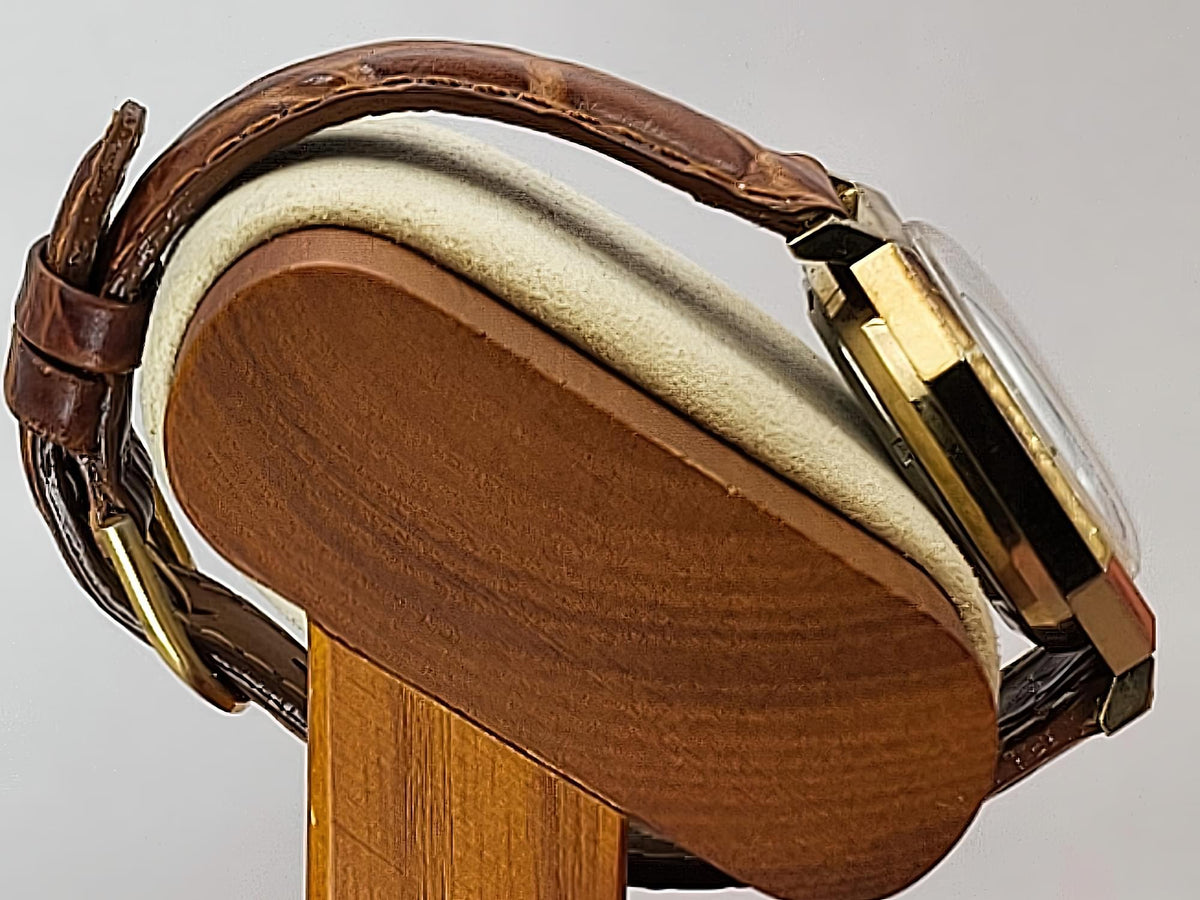 Close-up of a Caravelle Day-Date wristwatch with a brown leather strap and gold-tone case, displayed on a wooden stand against a light gray background.