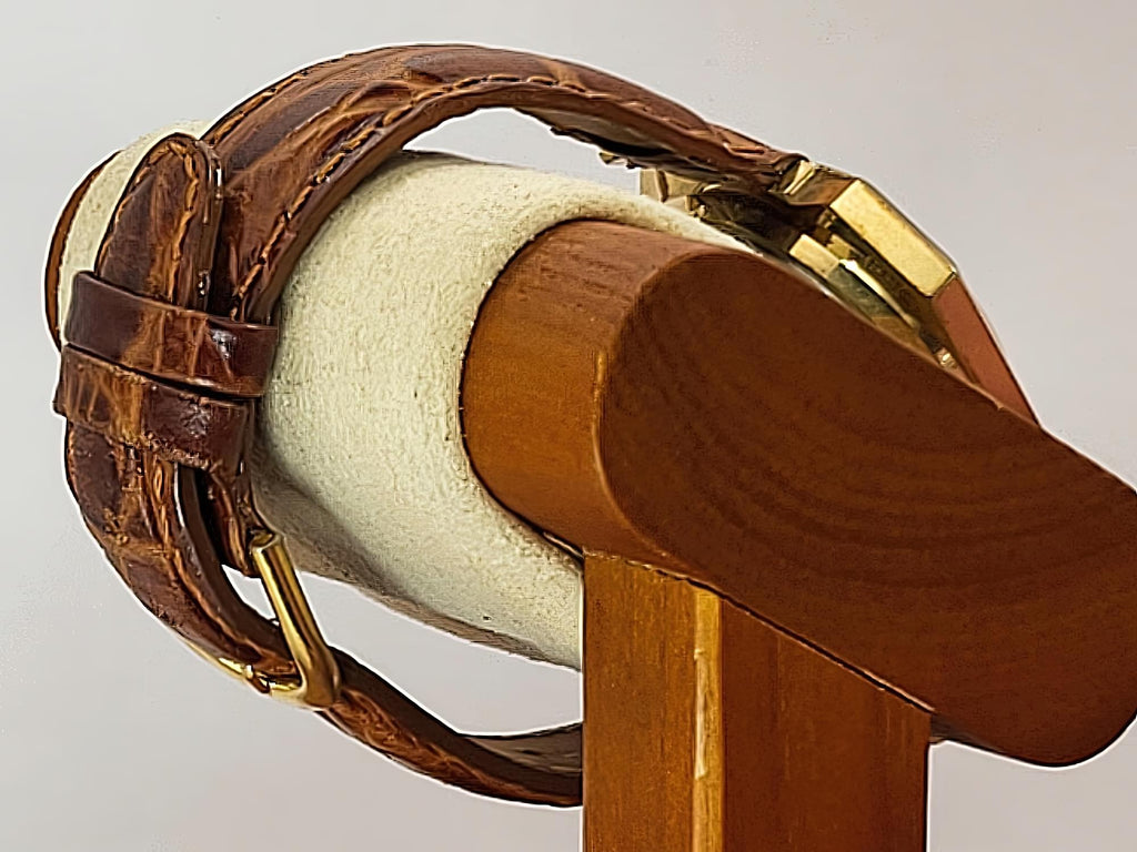 A close-up of a brown leather Caravelle Day-Date watch strap with gold-tone hardware, wrapped around a cream-colored display stand with a wooden base against a light gray background.