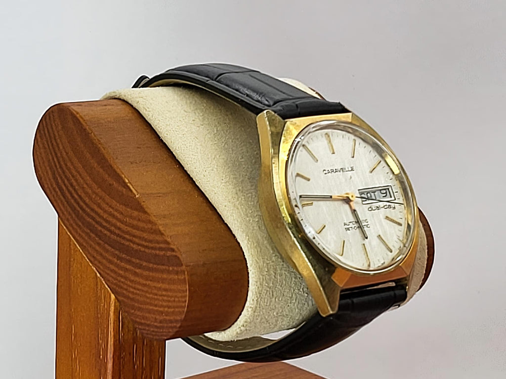 A vintage Caravelle Dual Day wristwatch with a gold-tone case, white dial, and black leather strap, displayed on a cream cushion mounted on a wooden stand against a light gray background.