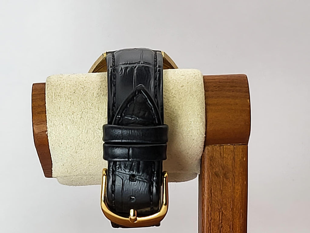 A black leather Caravelle Dual Day watch strap with a gold buckle, displayed on a wooden stand with a cream felt cushion against a light gray background.