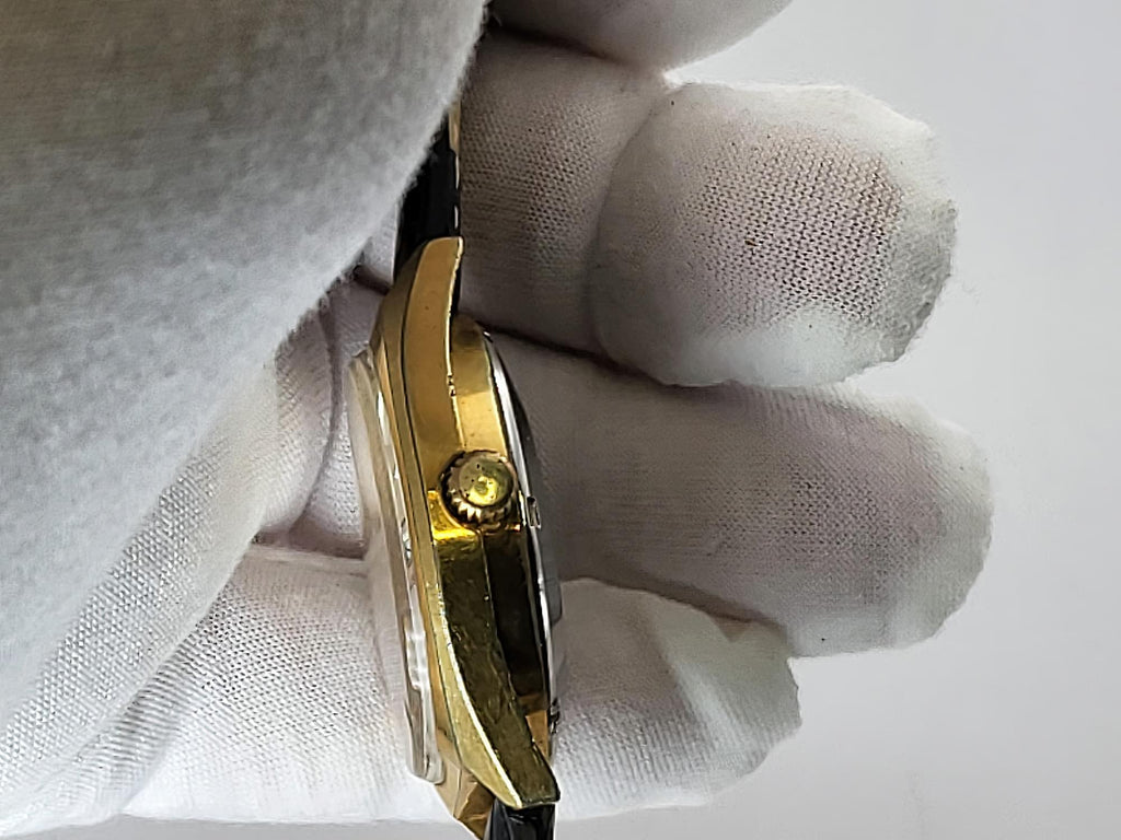 A close-up side view of a gold-tone Caravelle Dual Day wristwatch held by a hand wearing a white cotton glove, showing the side profile and crown against a plain light background.