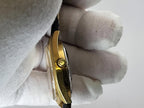 A close-up side view of a gold-tone Caravelle Dual Day wristwatch held by a hand wearing a white cotton glove, showing the side profile and crown against a plain light background.
