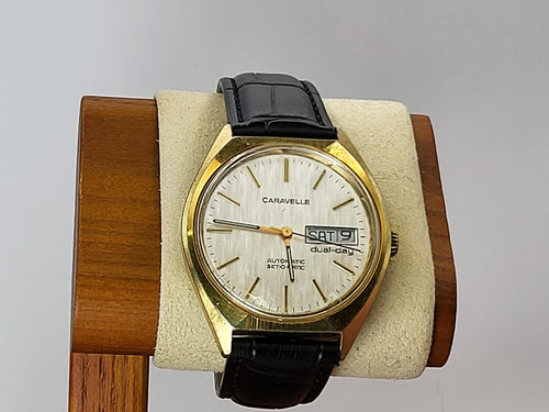 A vintage Caravelle Dual Day wristwatch with a gold-tone case, white dial, and black leather strap, displayed on a cream cushion mounted on a wooden stand.