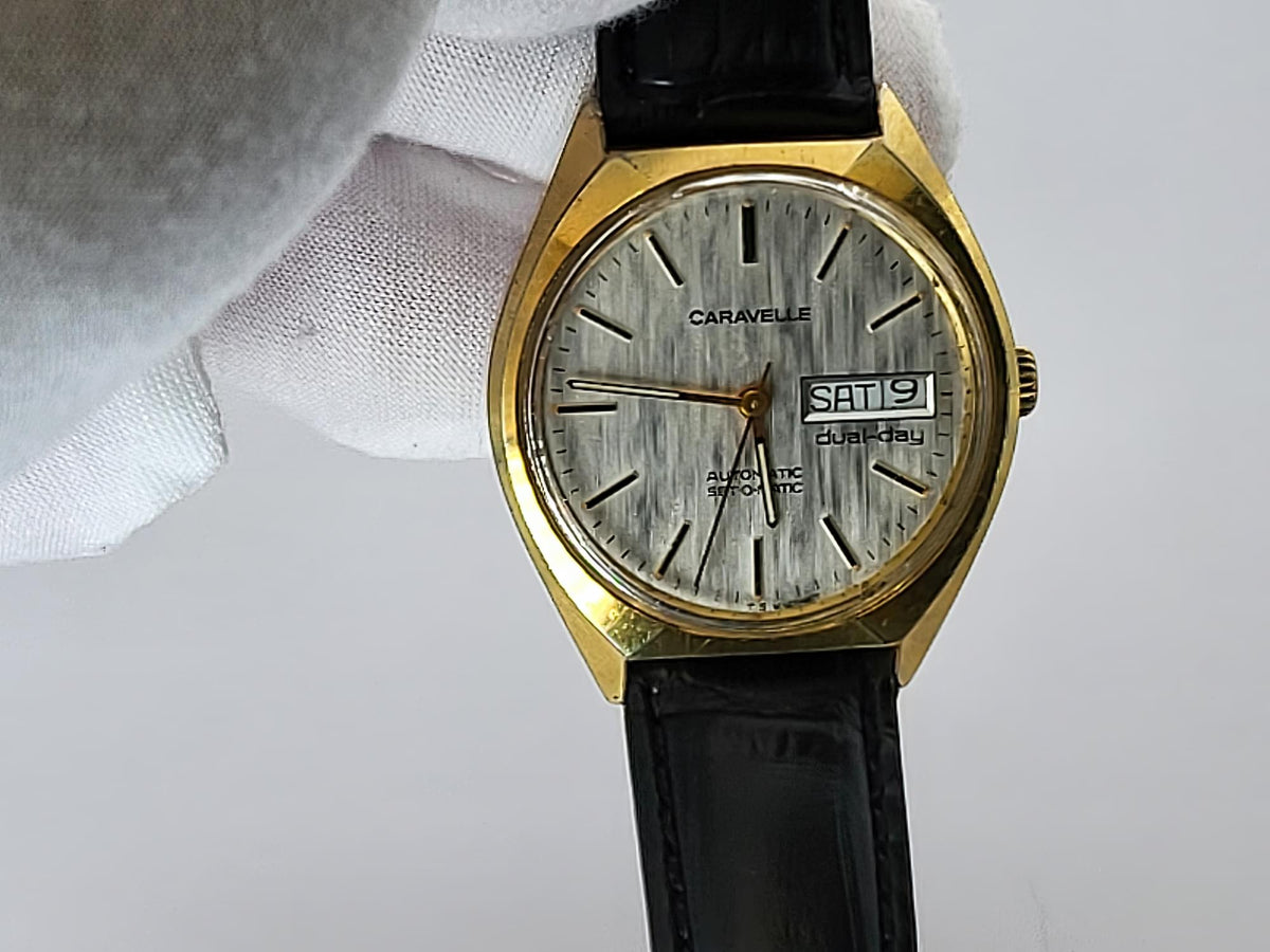 A vintage Caravelle wristwatch with a gold-tone case, silver dial, and black leather strap, held by a white gloved hand against a light gray background.