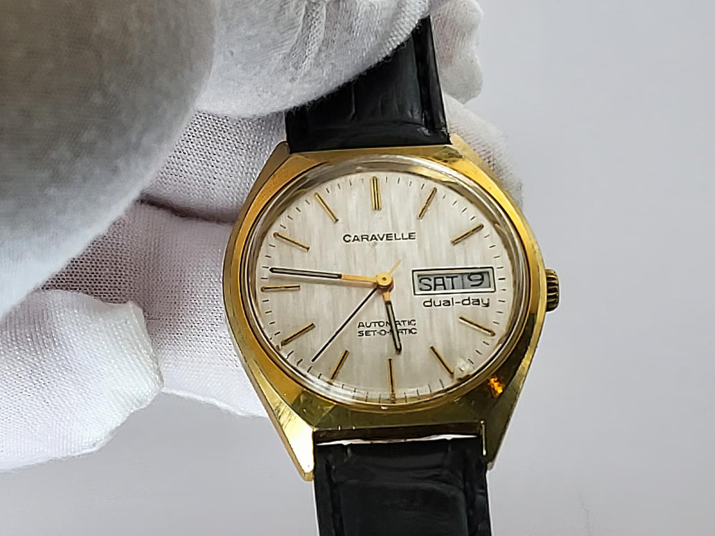 A vintage Caravelle dual-day automatic watch with a gold-tone case, cream-colored dial, and black leather strap, held by a white gloved hand against a light gray background.