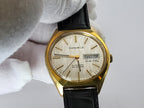 A vintage Caravelle dual-day automatic watch with a gold-tone case, cream-colored dial, and black leather strap, held by a white gloved hand against a light gray background.