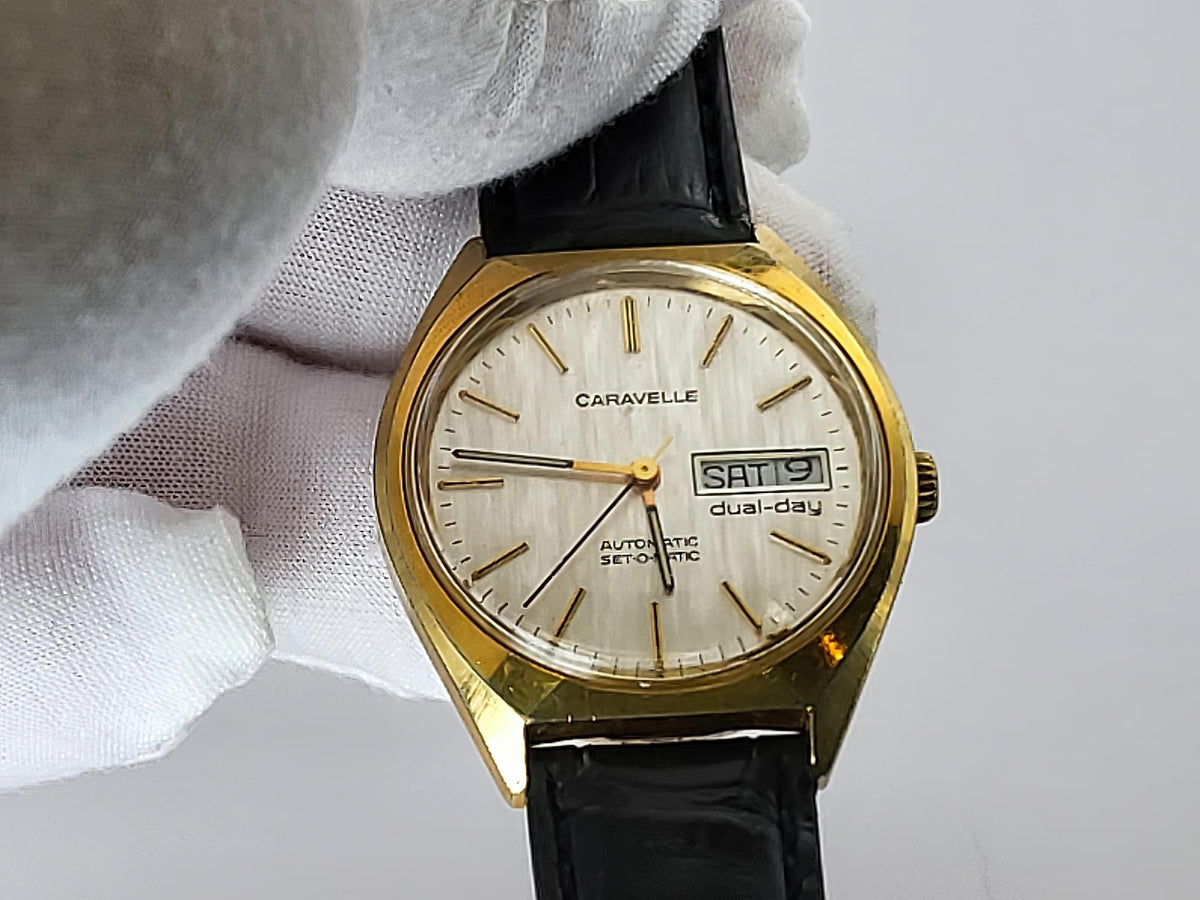A vintage Caravelle dual-day automatic watch with a gold-tone case, cream-colored dial, and black leather strap, held by a white gloved hand against a light gray background.
