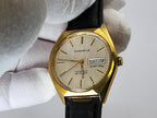 A vintage Caravelle wristwatch with a gold-tone case, cream dial, and black leather strap, held by a white-gloved hand against a light gray background.