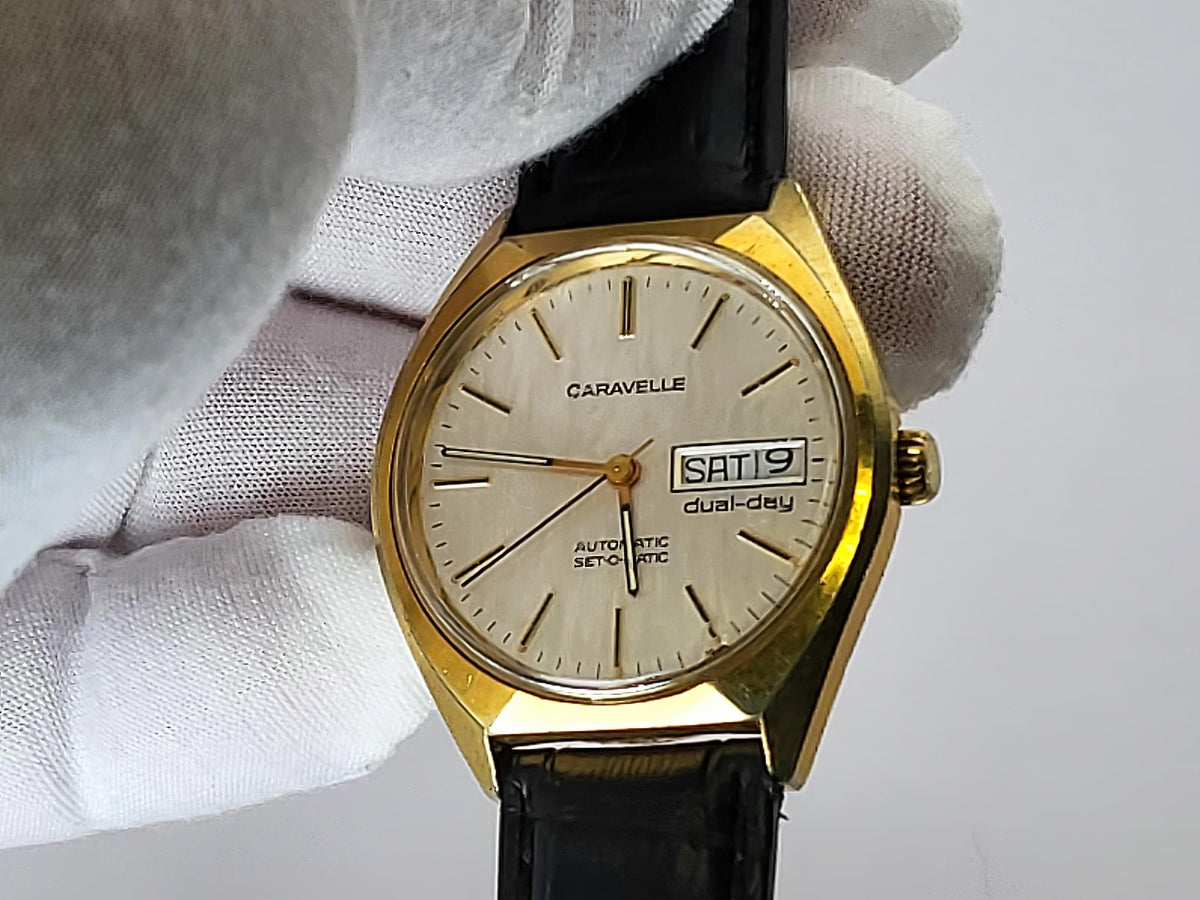 A vintage Caravelle wristwatch with a gold-tone case, cream dial, and black leather strap, held by a white-gloved hand against a light gray background.