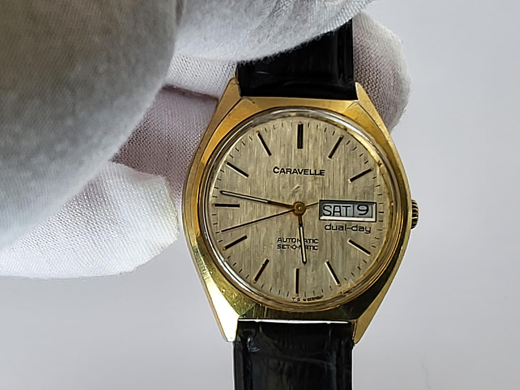 A vintage Caravelle dual-city watch with a gold-tone case, textured champagne dial, and black leather strap, held by a white-gloved hand against a light gray background.