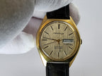 A vintage Caravelle dual-city watch with a gold-tone case, textured champagne dial, and black leather strap, held by a white-gloved hand against a light gray background.