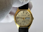 A vintage Caravelle dual-day wristwatch with a gold-tone case, cream dial, and black leather strap, held by a white-gloved hand against a light gray background.
