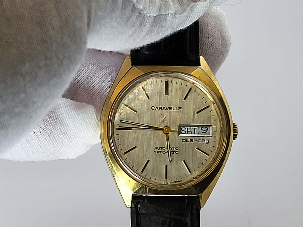 A vintage Caravelle dual-day wristwatch with a gold-tone case, cream dial, and black leather strap, held by a white-gloved hand against a light gray background.