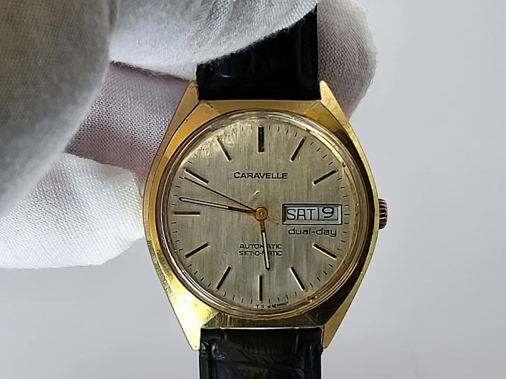 A vintage Caravelle automatic dual-day wristwatch with a gold-tone case, silver dial, and black leather strap, held by a white-gloved hand against a light gray background.