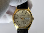 A vintage Caravelle automatic dual-day wristwatch with a gold-tone case, silver dial, and black leather strap, held by a white-gloved hand against a light gray background.