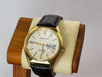A vintage  Caravelle Dual Day wristwatch with a gold-tone case, cream dial, and black leather strap, displayed on a beige cushion mounted on a wooden stand against a light gray background.