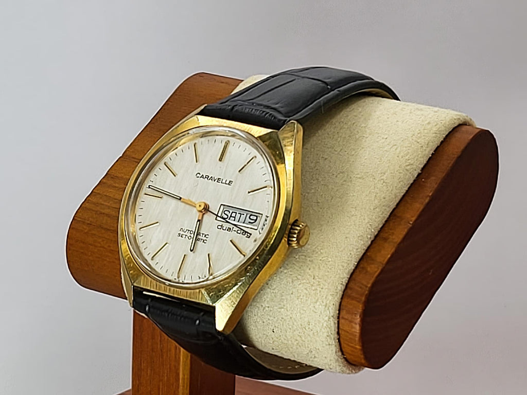 A vintage Caravelle Dual Day quartz watch with a gold-tone case, white dial, and black leather strap, displayed on a wooden stand with a cream cushion against a light gray background.