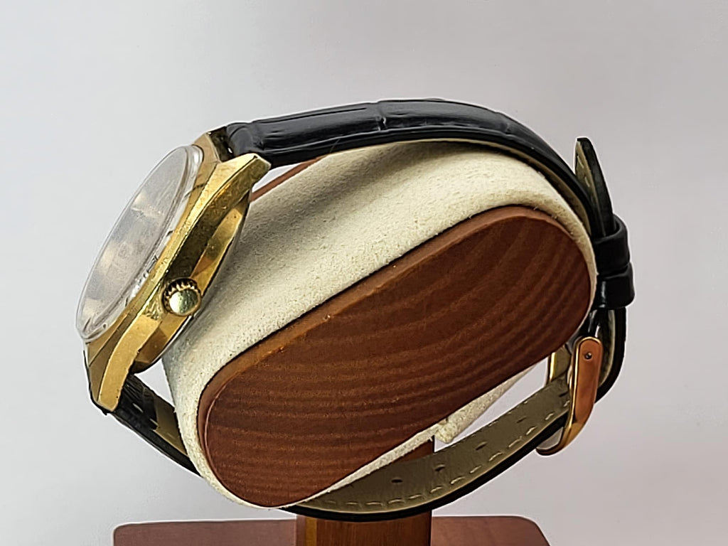 A vintage  Caravelle Dual Day wristwatch with a gold-tone case and black leather strap, displayed on a cream and wood cushion against a light gray background.