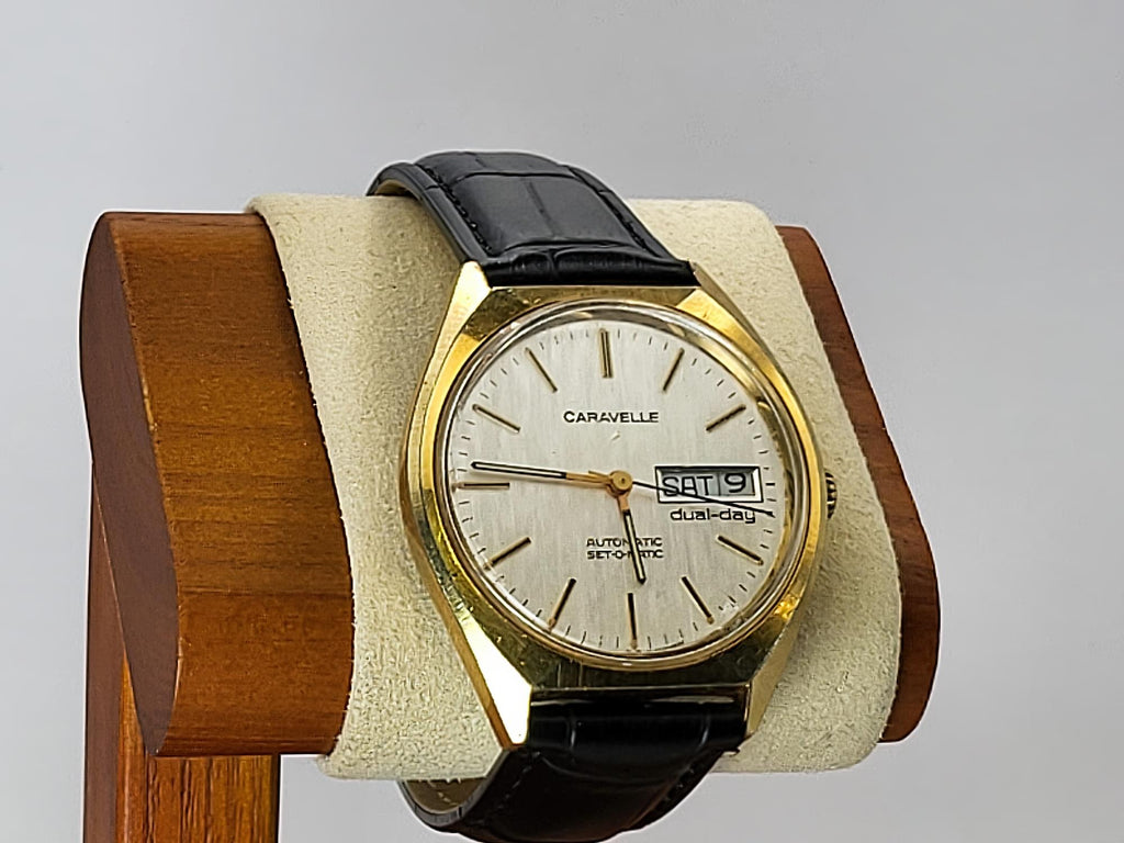 A vintage Caravelle wristwatch with a gold-tone case, cream-colored dial, and black leather strap, displayed on a beige cushion inside a wooden stand against a light gray background.