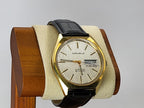 A vintage Caravelle wristwatch with a gold-tone case, cream-colored dial, and black leather strap, displayed on a beige cushion inside a wooden stand against a light gray background.