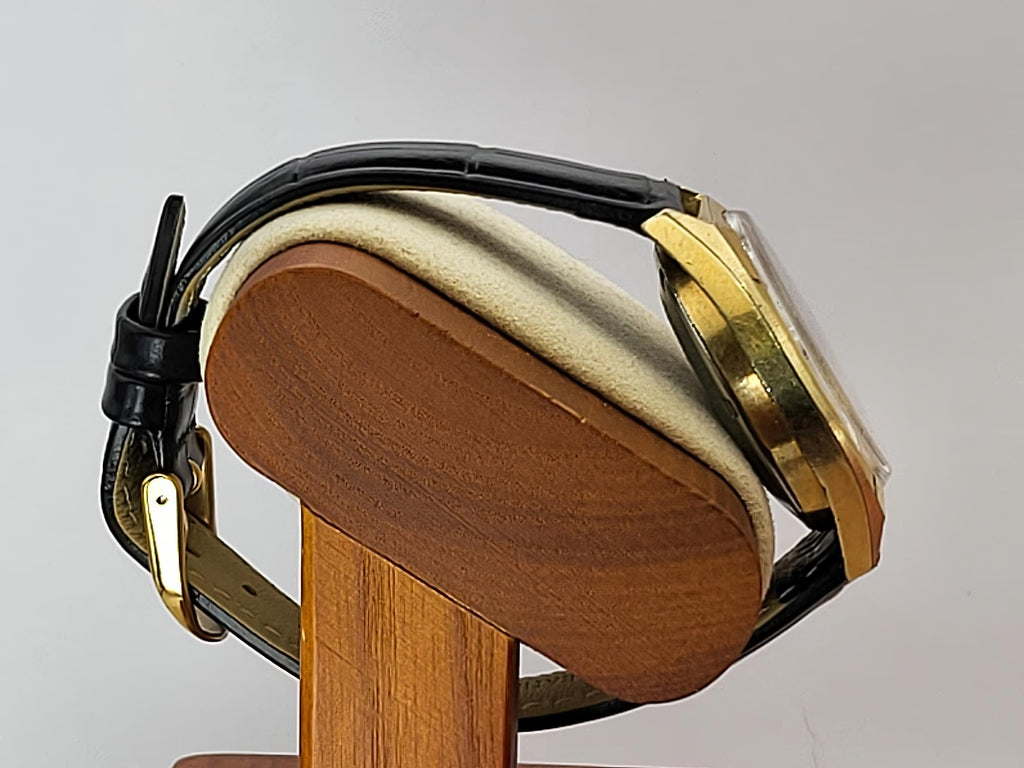 A vintage-style Caravelle Dual Day wristwatch with a black leather strap and gold-toned case, displayed on a wooden stand against a light gray background.