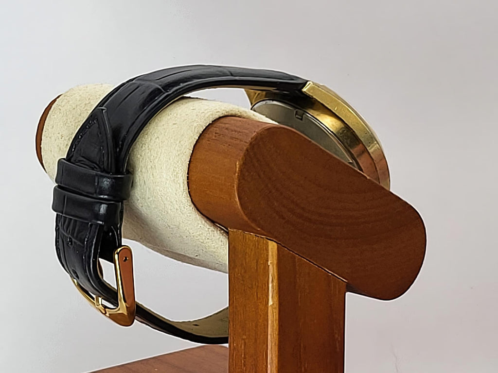 A close-up of a Caravelle Dual Day wristwatch with a black leather strap and gold-tone case, displayed on a wooden stand with a cream fabric roll.