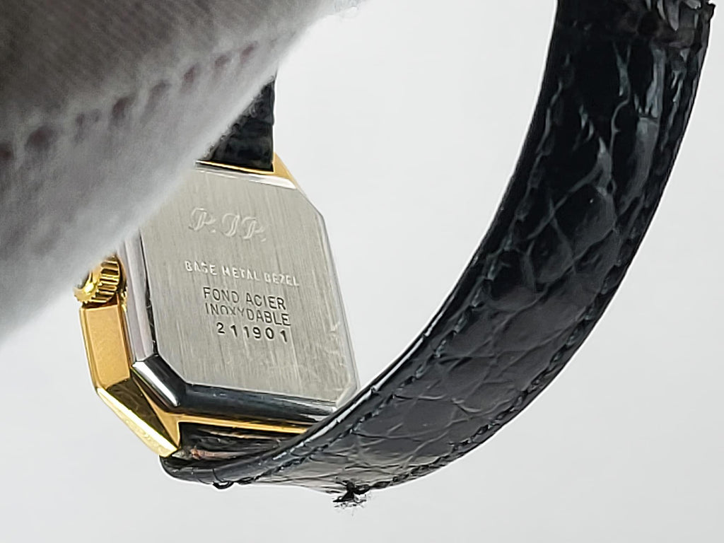 Close-up of the back of a Léonard Genève wristwatch with a gold-tone metal case, engraved silver-toned case back, and a black textured leather strap on a light gray background.