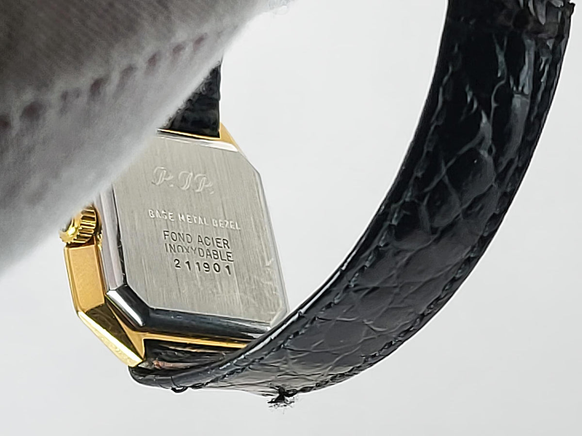 Close-up of the back of a Léonard Genève wristwatch with a gold-tone metal case, engraved silver-toned case back, and a black textured leather strap on a light gray background.