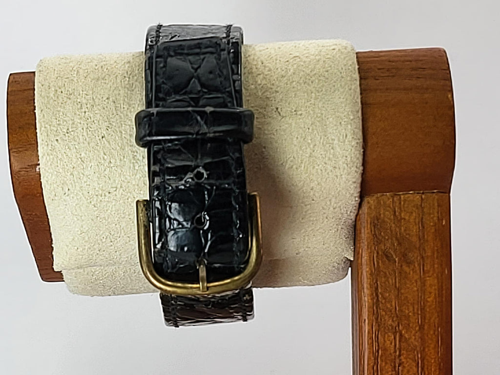 A black embossed leather watch strap with a gold-tone buckle for a Léonard Genève wristwatch, displayed on a cream-colored velvet watch stand set against a light gray background.