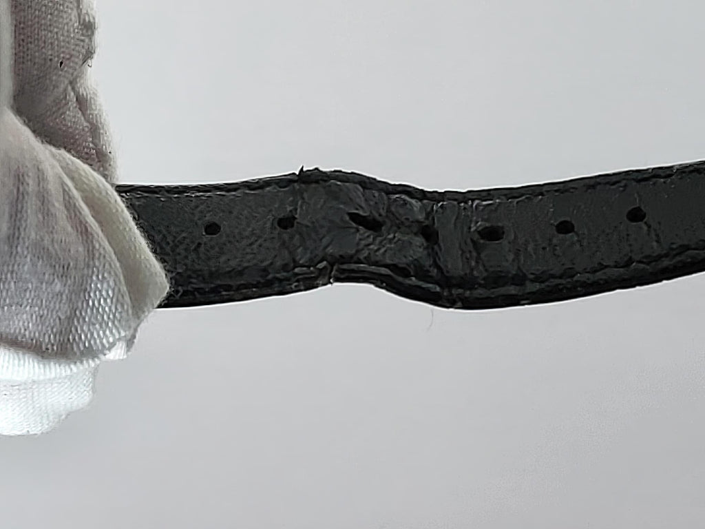 A close-up of a black leather belt strap for a Léonard Genève, held by a white gloved hand, showing a heavily worn, melted, or fused section in the center with missing or distorted holes.