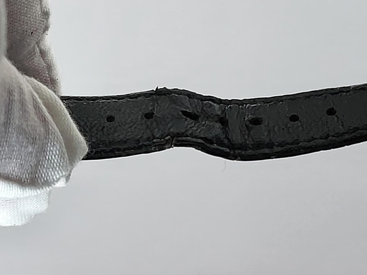 A close-up of a black leather belt strap for a Léonard Genève, held by a white gloved hand, showing a heavily worn, melted, or fused section in the center with missing or distorted holes.