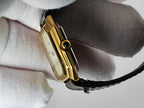 A close-up of a gold-tone rectangular Léonard Genève watch with a black strap, held by a hand wearing a white textured glove.