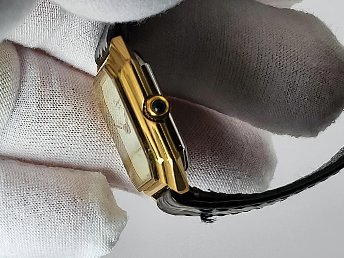 A close-up of a gold-tone rectangular Léonard Genève watch with a black strap, held by a hand wearing a white textured glove.
