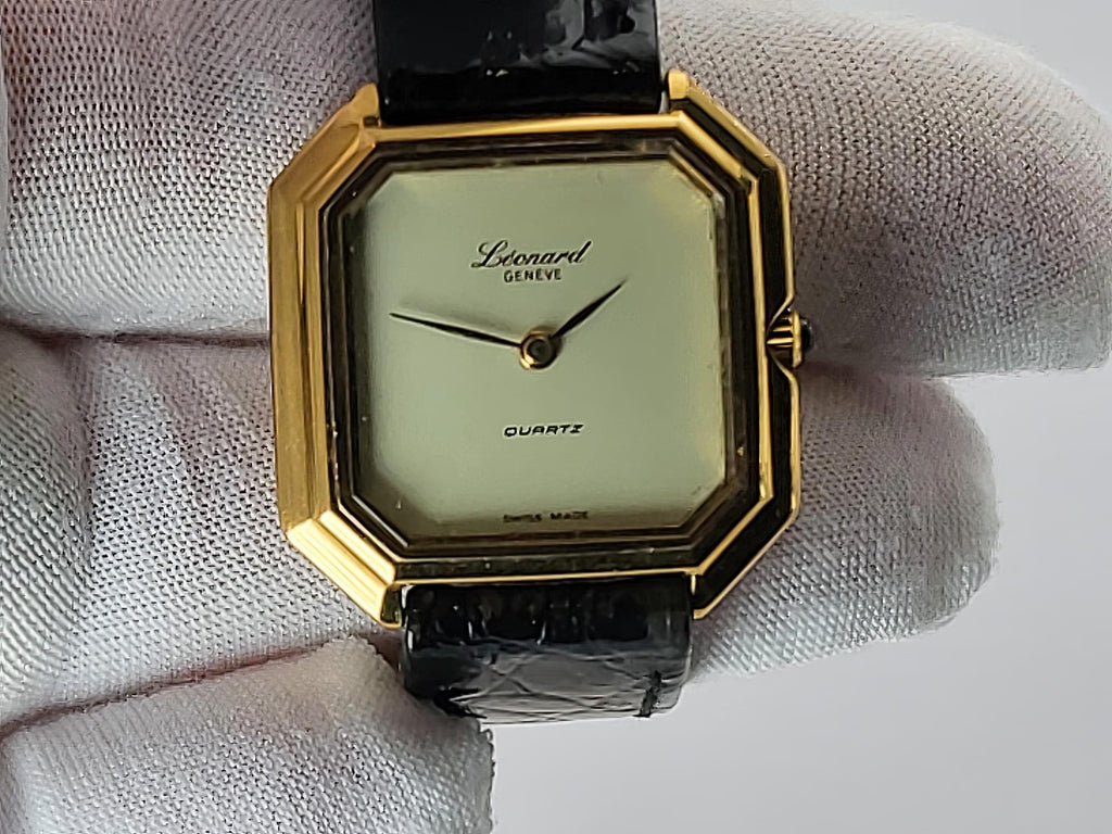 A vintage Léonard Genève quartz watch with an octagonal gold-tone case, cream-colored dial, and black leather strap, held by a white gloved hand.