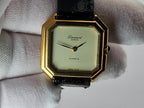 A Léonard Genève quartz watch with an octagonal gold-tone case, cream dial, and black strap, held in a white textured glove.