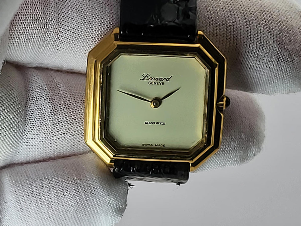 A Léonard Genève quartz watch with an octagonal gold-tone case, cream dial, and black strap, held in a white textured glove.