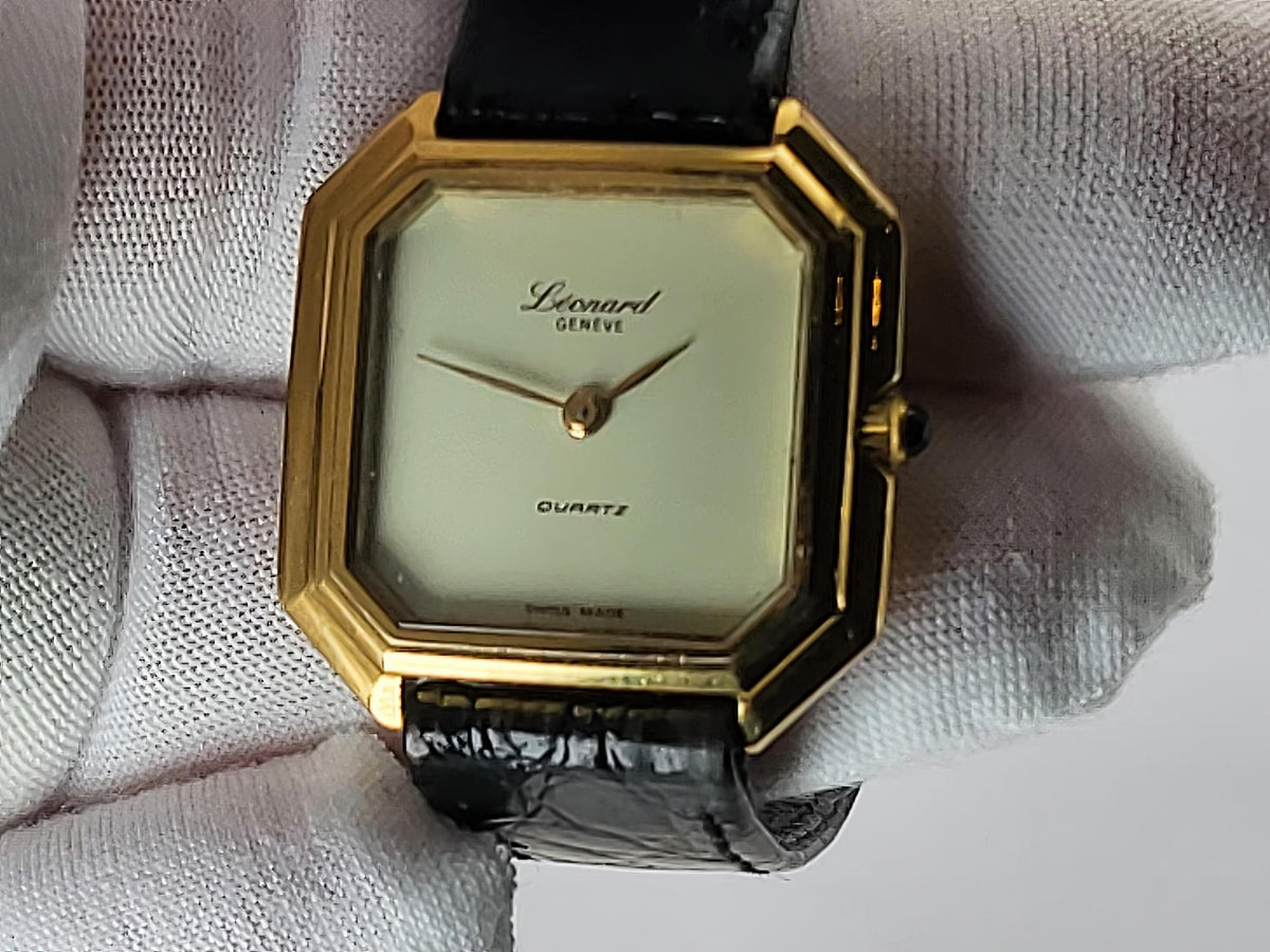 A vintage-style Léonard Genève wristwatch with an octagonal gold-tone case, cream dial, and black leather strap, shown resting on a white textured fabric surface.