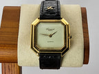 A vintage Léonard Genève watch with an octagonal gold-tone case, cream dial, and black leather strap, displayed on a beige cushion mounted on a wooden stand.