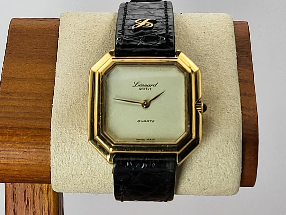 A vintage Léonard Genève watch with an octagonal gold-tone case, cream dial, and black leather strap, displayed on a beige cushion mounted on a wooden stand.