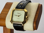 A vintage-style Léonard Genève wristwatch with an angular gold-tone case, cream dial, and black alligator-pattern strap, displayed on a beige cushion mounted on a wooden stand.