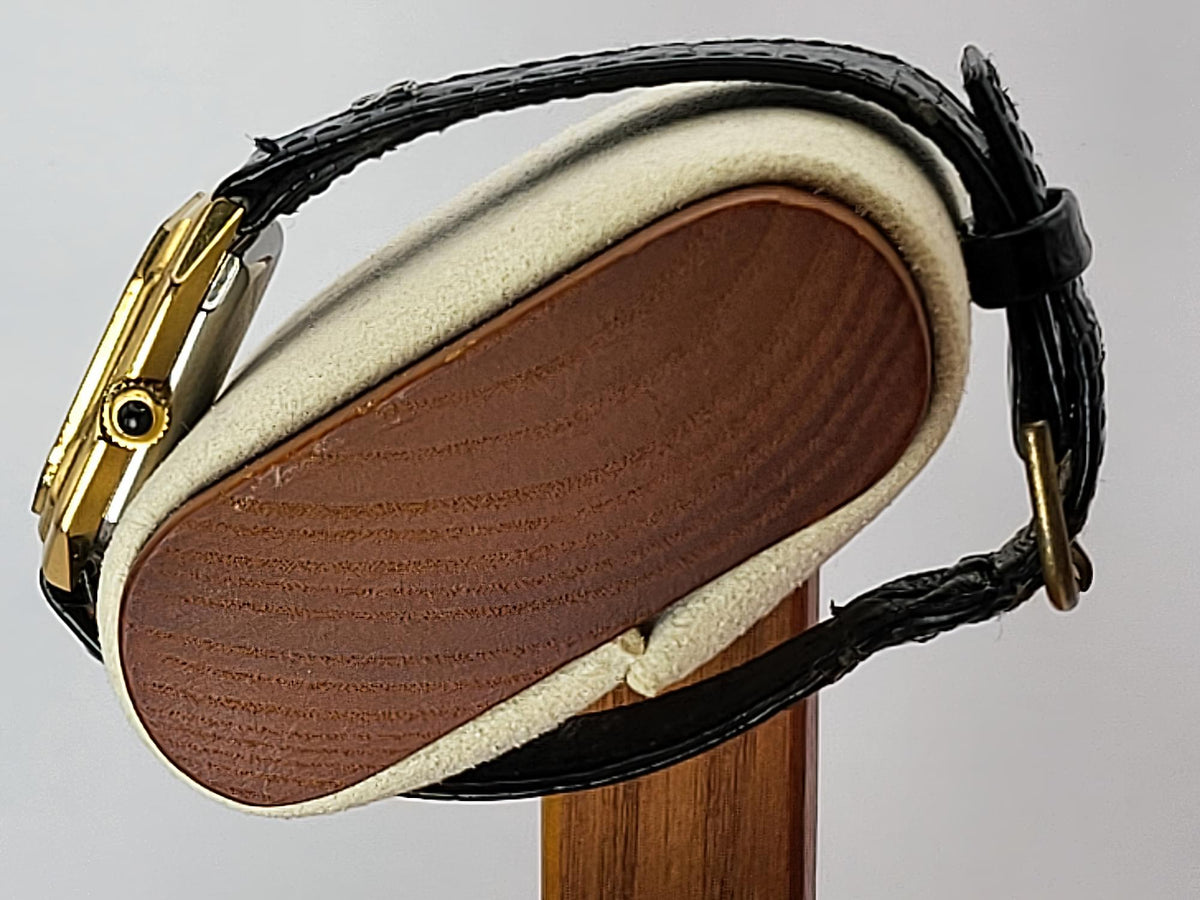 Close-up of a Léonard Genèvewristwatch strap and case on a display stand, showing a black textured leather strap, gold-toned metal case, and cream-colored cushioning around a brown inlay.