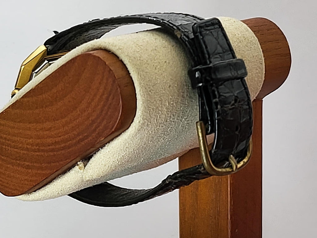 A black embossed leather watch strap with gold-tone hardware for a Léonard Genève wristwatch, displayed on a cream-colored wooden watch stand.
