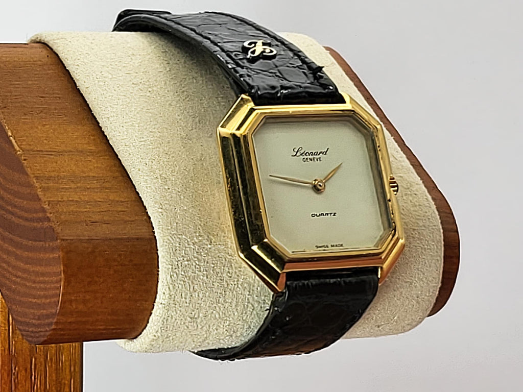 A vintage Léonard Genève quartz wristwatch with an octagonal gold-tone case, cream dial, and black alligator leather strap, displayed on a cream cushion mounted on a wooden stand.