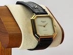 A vintage Léonard Genève quartz wristwatch with an octagonal gold-tone case, cream dial, and black alligator leather strap, displayed on a cream cushion mounted on a wooden stand.