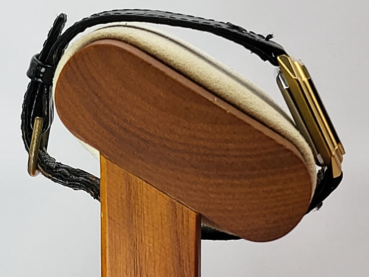 A close-up of a black leather watch strap with a gold buckle for a Léonard Genève watch, mounted on a wooden display stand against a light gray background.