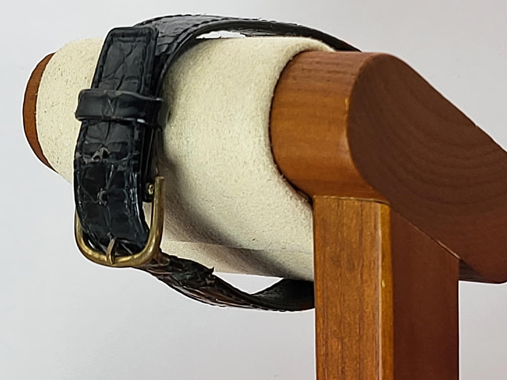 A close-up of a black embossed leather watch strap with a brass buckle for a Léonard Genève wristwatch displayed on a cream-colored cylindrical jewelry stand attached to a wooden holder.