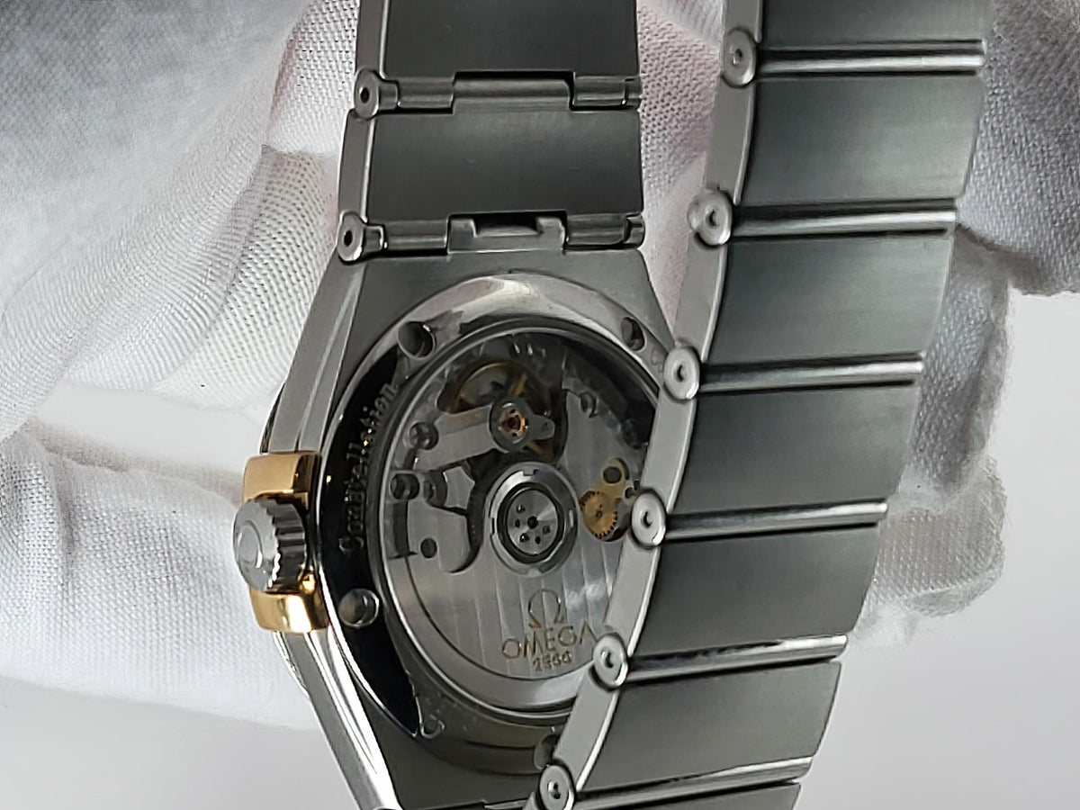Close-up of an Omega Constellation wristwatch back case with a transparent baffle showing the automatic movement, held against a white fabric background. The watch has a silver-toned metal bracelet and a gold-tone crown.